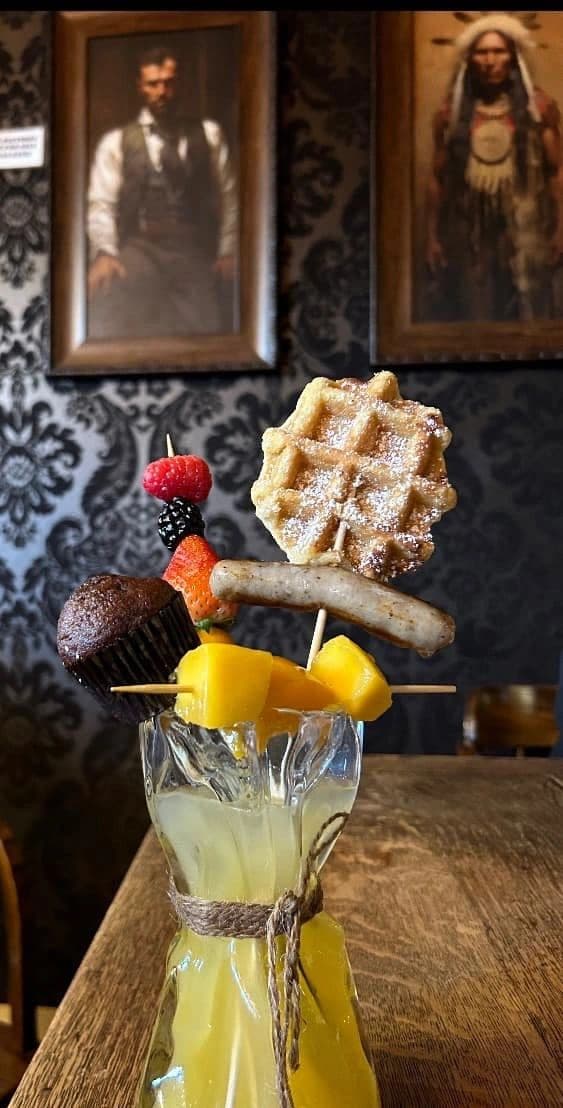 A signature fresh lemonade at the Smoke Wagon Saloon, served with a skewer of waffle, sausage, fruit, and a chocolate muffin, beneath framed Old West portraits.