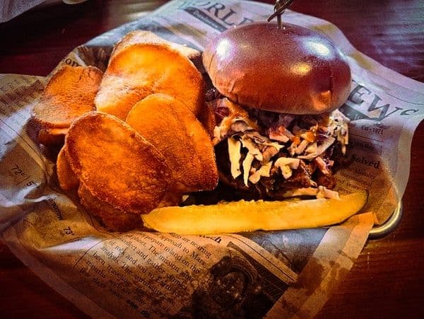 A smoked-meat sandwich with apple coleslaw on a brioche bun, served with house-made chips and a pickle.